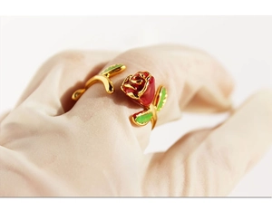 Detail of a handmade gold rose ring with red enamel and green leaves on a beige glove