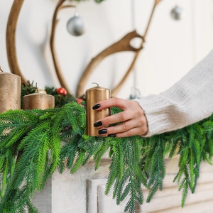 Golden pillar candle placed by hand on festive mantel with green Norfolk pine branches