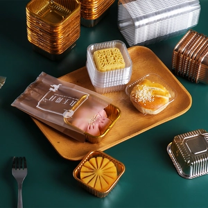 Golden and silver square mooncake trays with clear plastic lids on a dark table