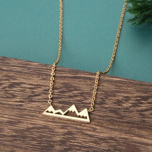 Close-up of golden mountain range hollow necklace on textured dark wood with pine needles
