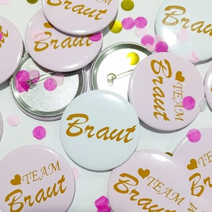 Flat lay of pink and white Braut tinplate badges with gold lettering for bridal parties