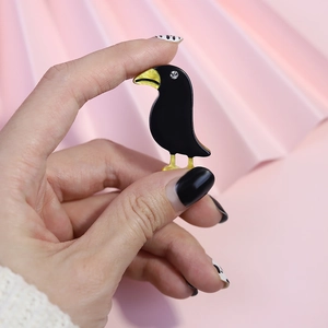 Fashionable black acrylic crow brooch with gold beak held by hand on pink background