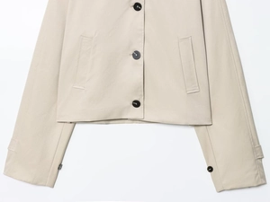 Dettaglio di trench corto beige con bottoni in resina scura e tasche laterali simmetriche