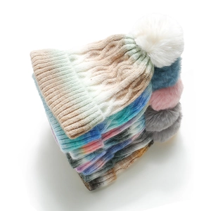 Detail of stacked tie-dye knit beanies featuring large faux fur pom-poms and thick texture