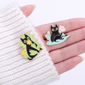 Hand holding two cute cartoon black cat enamel pins with floral motifs on a white sweater