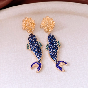 Detail of fish tassel earring with blue enamel scales and gold flower stud on beige tray