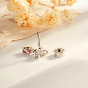 Silver bow and red heart pendant cartilage stud earrings on a white surface