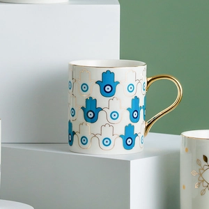 White ceramic mug with gold handle and blue Hamsa Evil Eye pattern in a modern setup