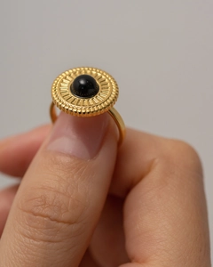 Dettaglio di un anello a disco dorato con gemma nera e trama a raggiera tenuto in mano