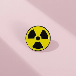 Close-up of a yellow and black radioactive symbol enamel pin on a soft pink background