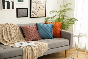 Three velvet diamond quilted pillows in pink, blue and orange with pompom fringe on a sofa