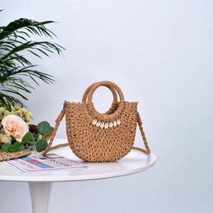 Handmade straw half-moon crossbody bag with seashell decor on a stylish white table