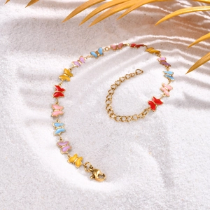 Detailed view of gold butterfly anklet with multi-colored enamel on beach sand