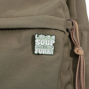 Close-up of enamel pin with Life is a Soup and I am a Fork quote on green canvas bag