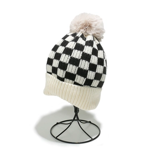 Detail of checkered knit beanie featuring a beige faux fur pompom and ribbed cuff