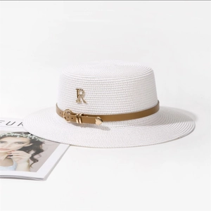 Détail d'un chapeau de paille blanc avec bande en cuir marron et breloque dorée avec lettre R