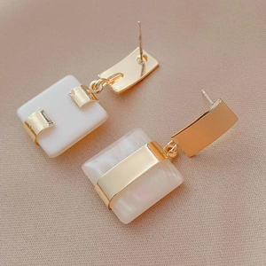 Détail de boucles d'oreilles dorées avec pendentifs carrés en nacre sur un tissu beige