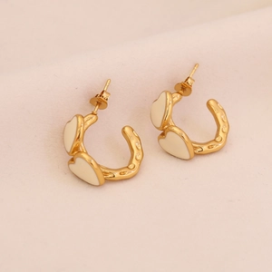 Macro detail of C-shaped gold earrings with white heart charms and hammered metal texture
