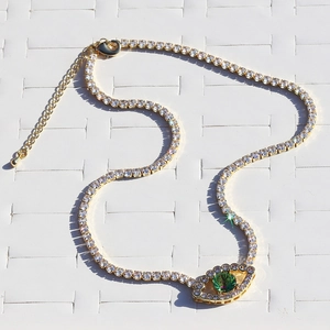 Detailed close-up of gold tennis necklace with green zircon eye pendant on grid mat