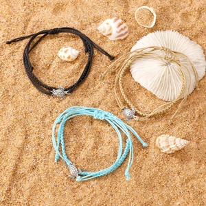 Three handmade woven ankle bracelets with silver turtle charms displayed on beach sand