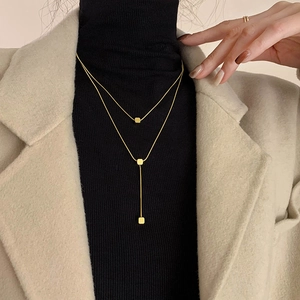 Double-layered gold necklace with square pendants worn over black turtleneck and beige suit