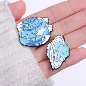 Hand holding two cute blue cat planet enamel pins on a soft white knitted sweater background