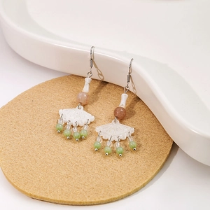 Detailed shot of white fan-shaped drop earrings with green beaded tassels on cork mat