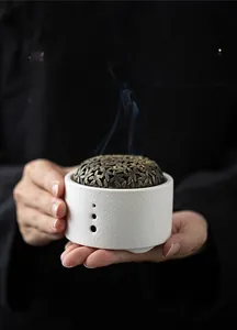 Close-up of hands holding a white ceramic incense burner with a bronze maple leaf lid and smoke