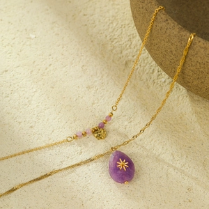 Double-layered gold necklace with purple teardrop stone and heart charm on stone background