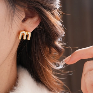 Close-up of golden letter M stud earring with white enamel inlay on female model ear