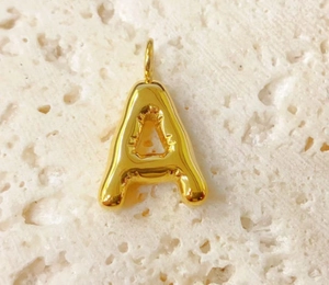 Detailed view of a 3D bubble letter A pendant in gold finish on a beige stone surface