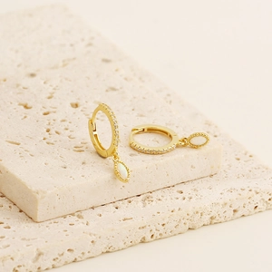 Close-up of gold hoop earrings with white opal drops and pavé crystals on stone pedestal