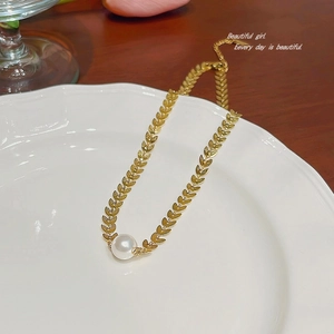 Close-up of golden wheat link necklace with a white pearl on a white ceramic plate