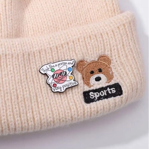 Close-up of beige knit hat featuring a proton enamel pin and bear head embroidery