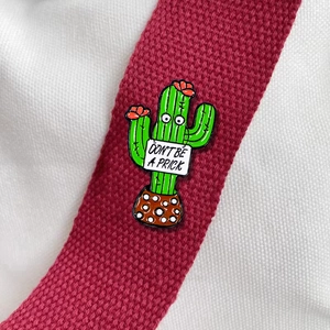 Cartoon cactus enamel pin with Don't Be A Prick sign on burgundy fabric