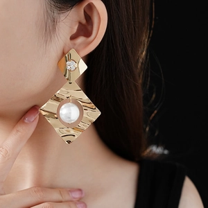 Detail of a large gold wavy metal earring featuring a rhombus design with a white pearl