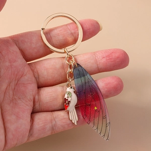 Golden bird keychain with rainbow wing pendant shown held by hand against beige background