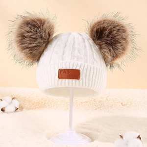 Detail of white knit baby beanie with double brown faux fur pom poms and leather label