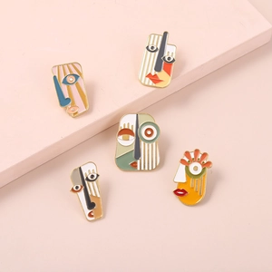 Close-up of five cubist enamel face pins displayed on modern pink geometric stages