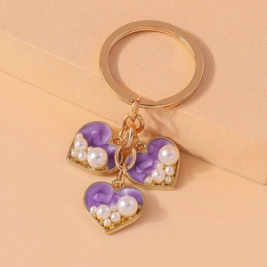 Close-up of golden heart keychain with purple enamel and embedded pearls on beige background