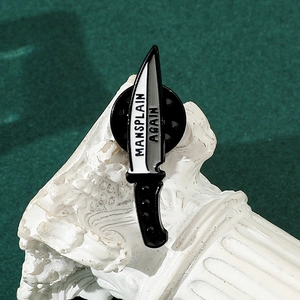 Close-up of enamel dagger brooch with MANSPLAIN AGAIN text on white plaster prop