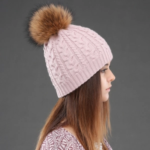 Close-up of pink cable knit wool hat featuring a fluffy raccoon fur pom-pom detail