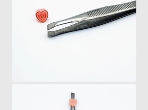 Macro shot of tweezers holding a small pink heart charm featuring an engraved shield and M letter