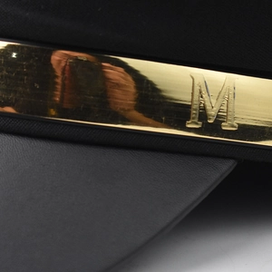 Detail of black newsboy hat featuring a leather brim and gold metal bar with M engraving