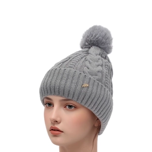 Détail du bonnet en tricot gris avec motif torsadé, pompon moelleux et bijou de feuille