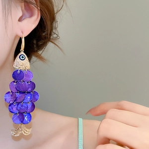 Detail of gold fish-shaped earring with purple sequins and evil eye charm on model