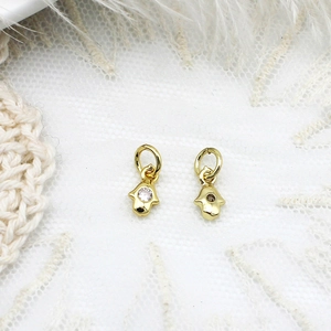 Macro detail of gold Hamsa hand charms with clear zircon on a soft lace background
