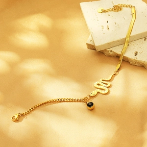 Detail of gold snake bone chain with evil eye and heart charms on a beige textured stone