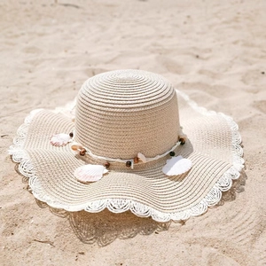 Close-up of wavy brim straw hat featuring decorative seashells and beads on sandy beach