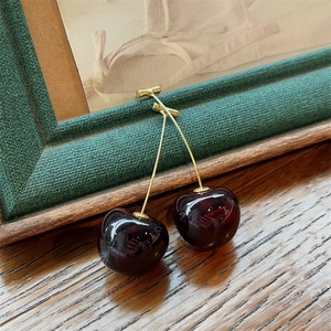 Close-up of deep red cherry earrings with gold stems resting on a green vintage frame